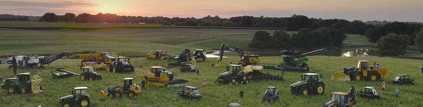 Ľudia kráčajúci medzi niekoľkými typmi zariadení John Deere na veľkom poli.