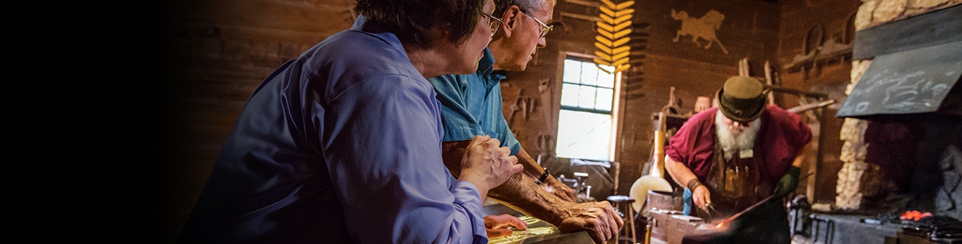 Starší pár sledujúci kováča pri práci s kovovým kladivom na historickej lokalite v Grand Detour, Illinois v Spojených štátoch.