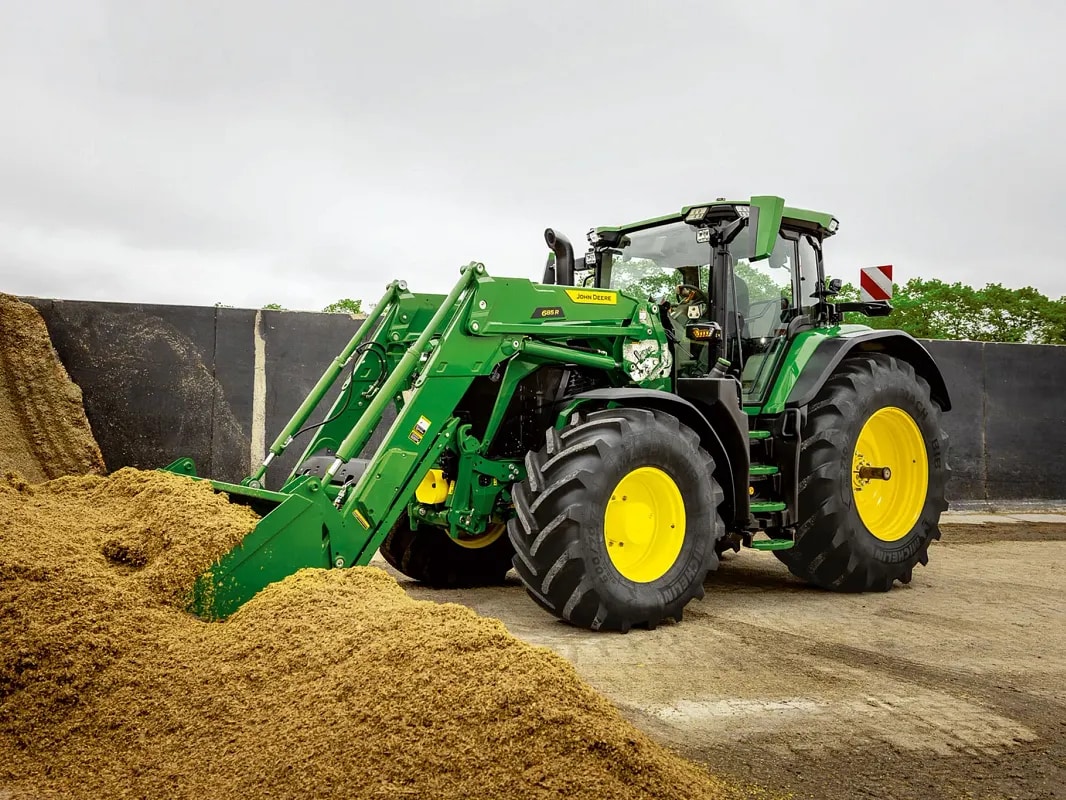 Traktor John Deere tlačí pomocou lyžice čelného nakladača do silážnej hromady v prostredí farmy.
