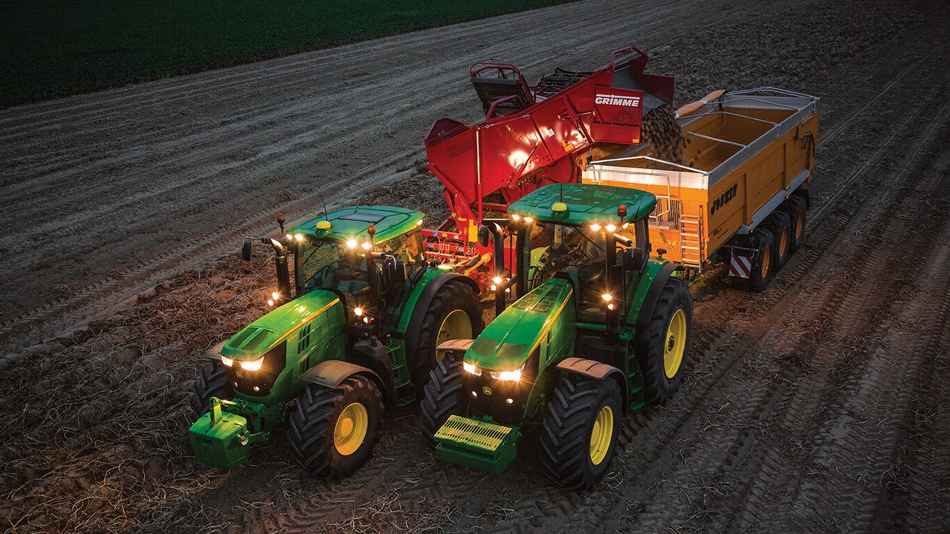 Traktor ťahaj&uacute;ci stroj na zber zemiakov v noci vyklad&aacute; zemiaky na vozidlo, ktor&eacute; ťah&aacute; in&yacute; traktor
