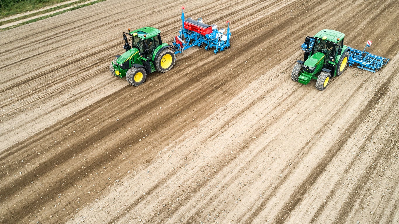 Dva traktory John Deere 6M na holom poli. Jeden ťah&aacute; sejačku Monsomen.  Druh&yacute; ťah&aacute; sejaciu kombin&aacute;ciu Lemken