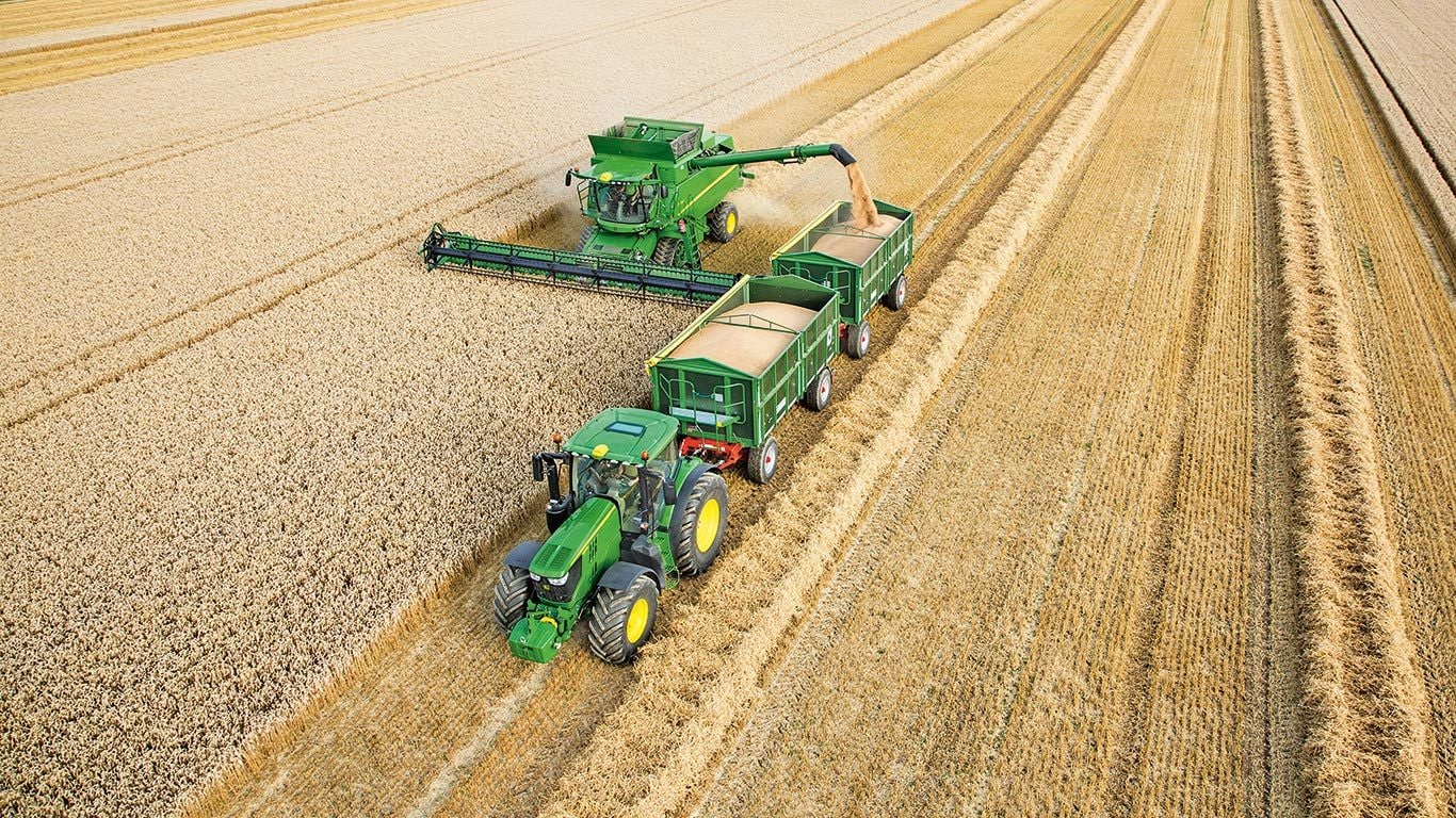 Kombajn John Deere radu S vyklad&aacute; p&scaron;enicu do du&aacute;lnych pr&iacute;vesov, ktor&eacute; ťah&aacute; traktor