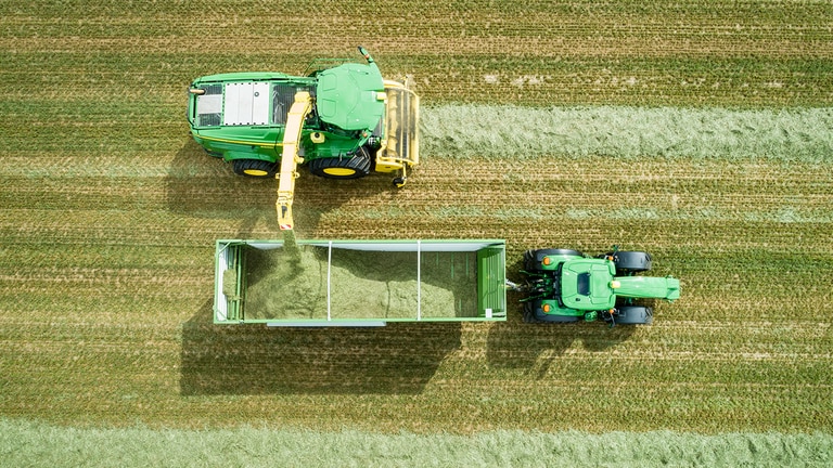 Leteck&aacute; sn&iacute;mka samohybn&eacute;ho kombajnu John Deere radu 8000, ktor&yacute; vyklad&aacute; sil&aacute;ž do vozidla, ktor&eacute; ťah&aacute; traktor John Deere