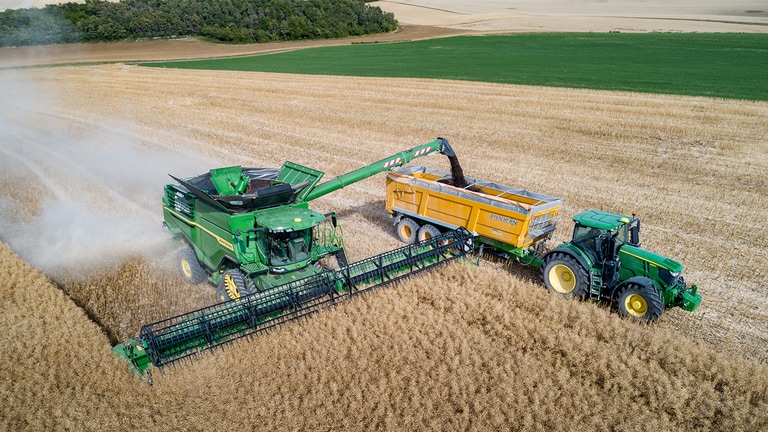 Kombajn John Deere radu X vyklad&aacute; repku do vozidla na obilie, ktor&eacute; ťah&aacute; traktor John Deere