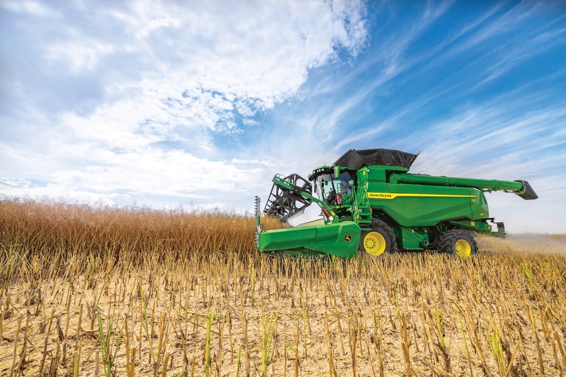 Kombajn John Deere žne obilie so žacím valom na poli pod modrou oblohou počas zberu plodiny