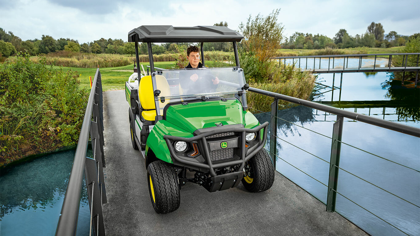 Úžitkové vozidlo John Deere Gator GS prechádzajúce úzkym mostom ponad pokojnú vodnú plochu, obklopené bujnou zeleňou a stromami. Úžitkové vozidlo John Deere Gator GS prechádzajúce úzkym mostom ponad pokojnú vodnú plochu, obklopené bujnou zeleňou a stromami.
