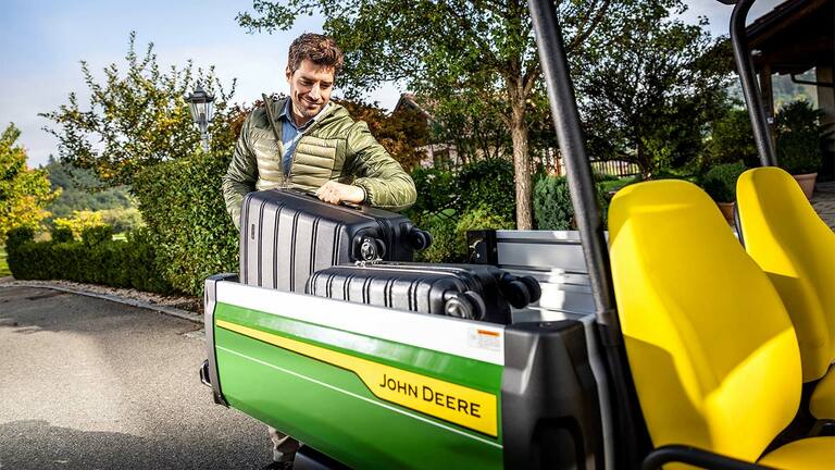 Osoba nakladajúca veľký čierny kufor do nákladového priestoru vozidla John Deere Gator GS s jasne žltými sedadlami, obklopený zeleňou a stromami.