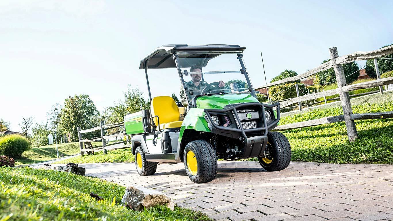 Zelené úžitkové vozidlo John Deere Gator GS jazdiace po dláždenej ceste cez upravenú oblasť s trávou a drevenými plotmi.