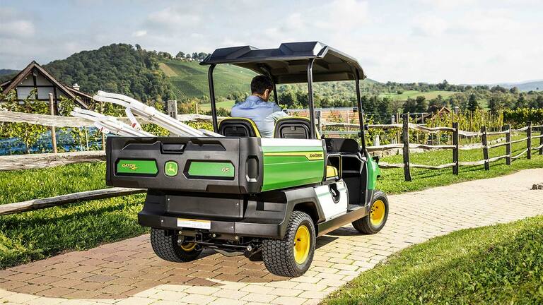 Zadný pohľad na vozidlo John Deere Gator GS jazdiace po ceste z tehál s malebnými kopcami a vinicami v pozadí.