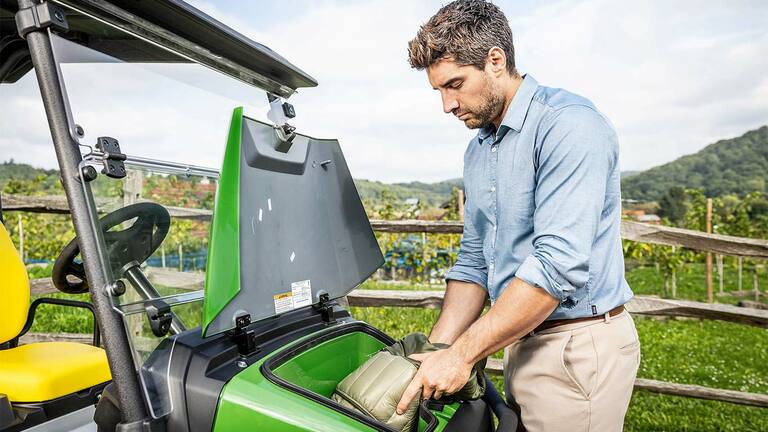 Osoba vkladajúca vak do predného úložného priestoru zeleného vozidla John Deere Gator GS zaparkovaného vonku pri drevenom plote a vinici.
