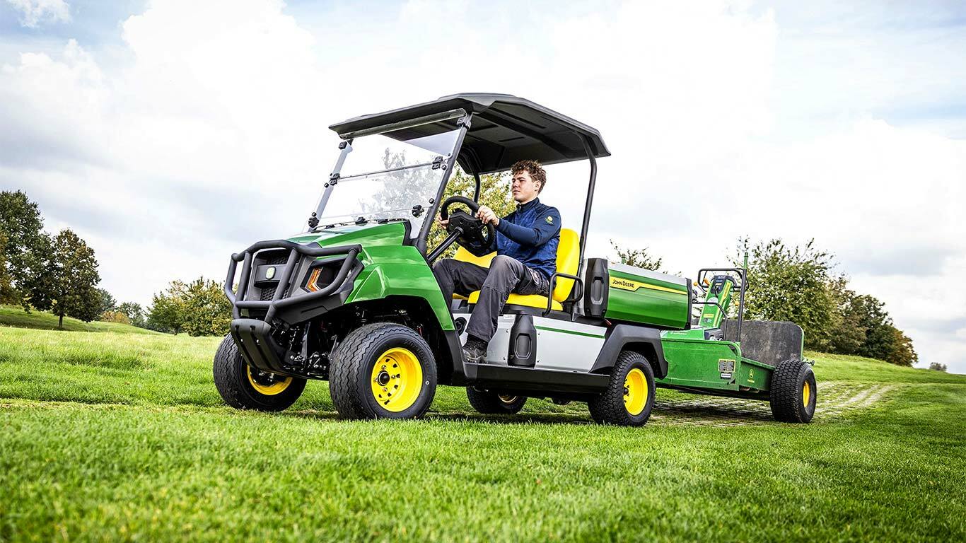 Zelené elektrické úžitkové vozidlo John Deere Gator ťahajúce vybavenie cez trávnaté pole pod čiastočne zamračenou oblohou.