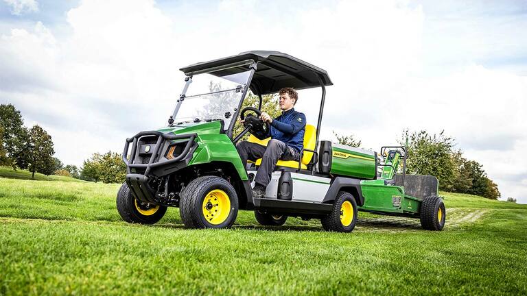 Zelené elektrické úžitkové vozidlo John Deere Gator ťahajúce vybavenie cez trávnaté pole pod čiastočne zamračenou oblohou.