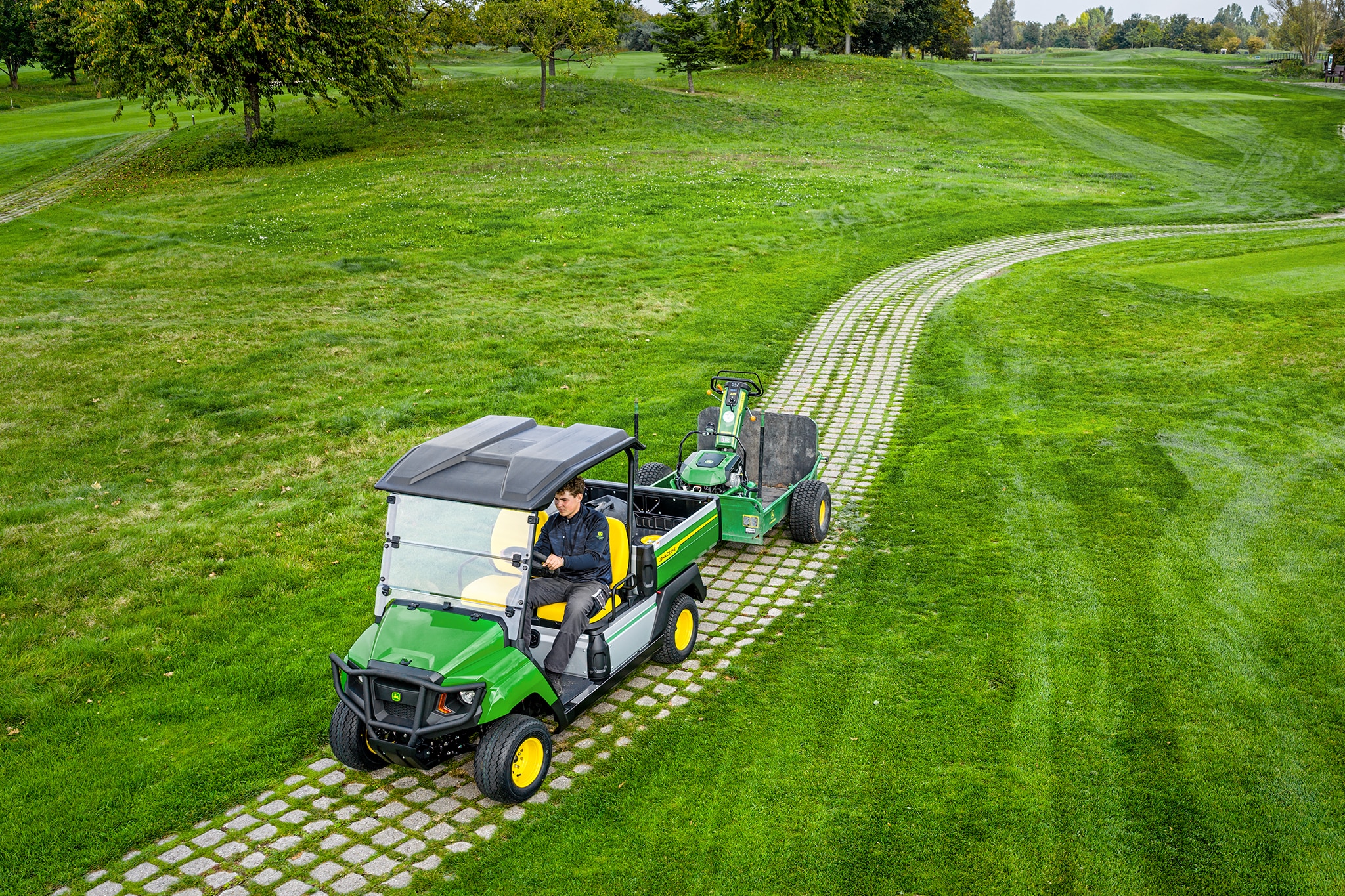 John Deere Gator GS ťahajúci vybavenie po trávnatej cestičke na golfovom ihrisku John Deere Gator GS ťahajúci vybavenie po trávnatej cestičke na golfovom ihrisku