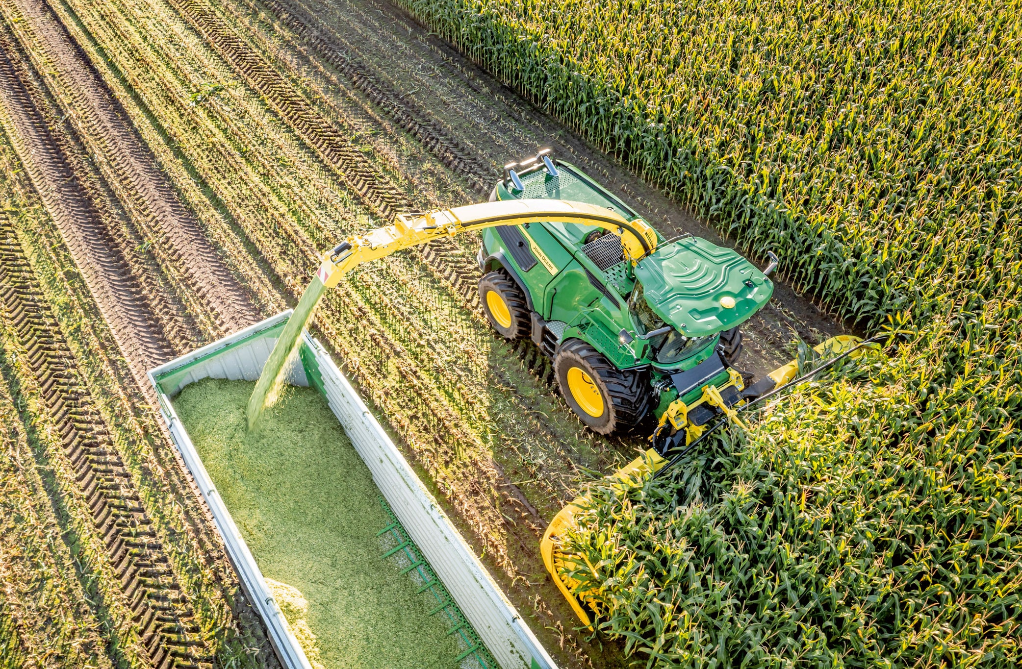 Letecký pohľad na zelený kombajn John Deere, ktorý na veľkom kukuričnom poli zberá kukuricu a prekladá nasekanú siláž do prívesu.