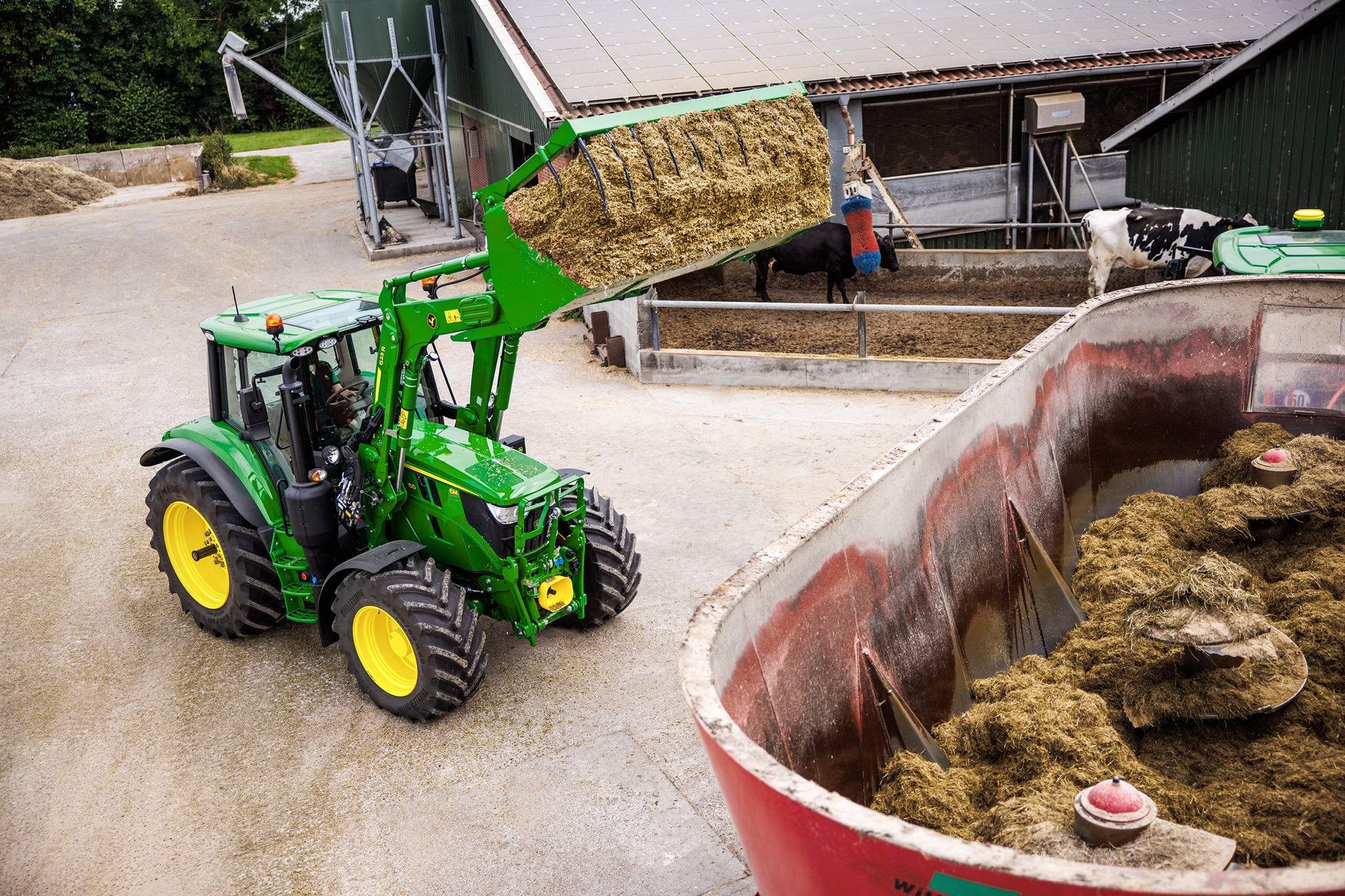 Traktor John Deere radu 6M používajúci čelný nakladač na nakladanie siláže do prívesného zmiešavača na dvore farmy.