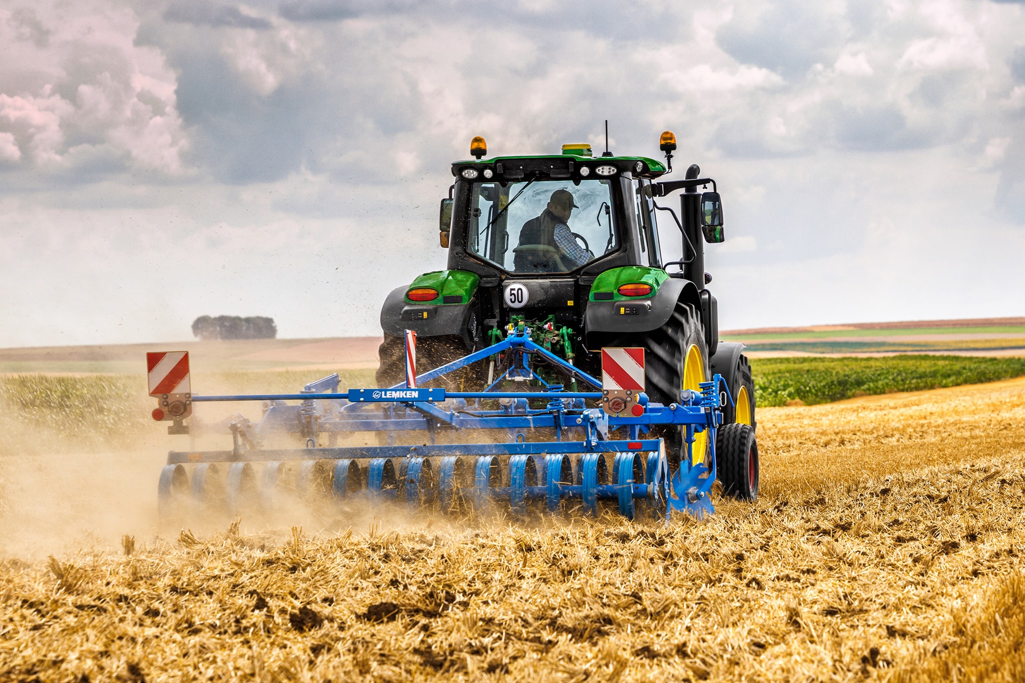 Traktor John Deere radu 6M obrába zožaté pole s diskovými bránami, pohľad zozadu v prašných poľných podmienkach.