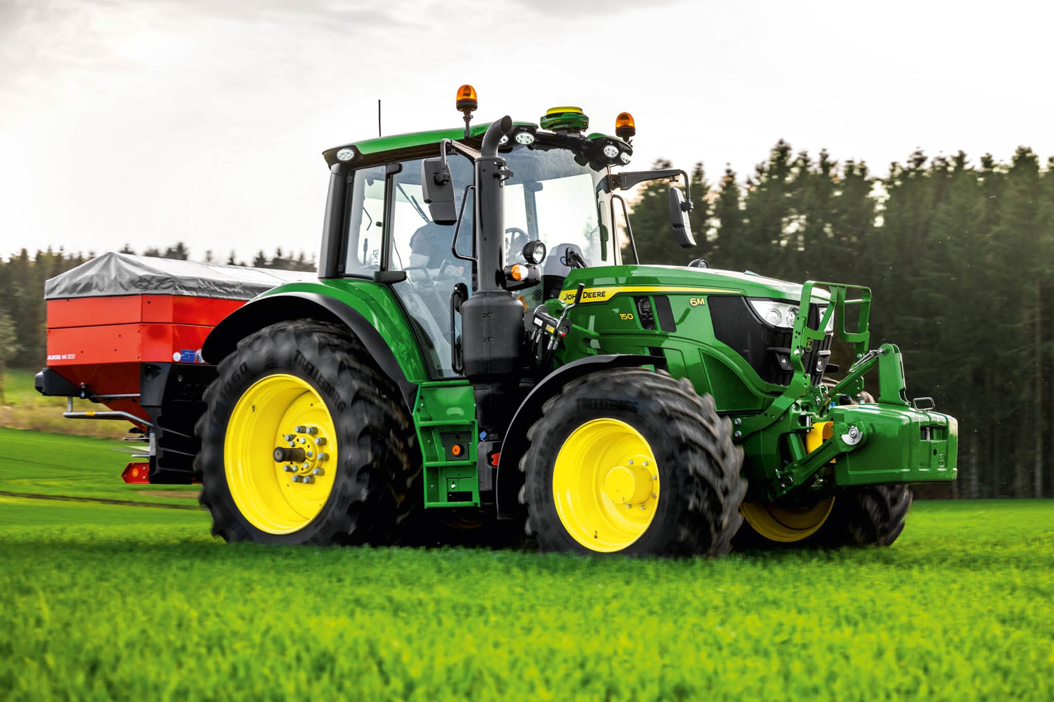 Traktor John Deere radu 6M so zadným rozmetadlom pri práci na tráve v blízkosti okraja lesa.
