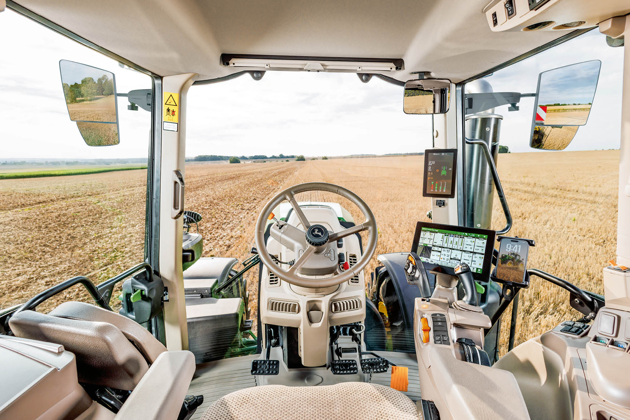 Interiér kabíny traktora John Deere s volantom, digitálnymi ovládacími displejmi a výhľadom na pole cez čelné sklo počas práce na farme.