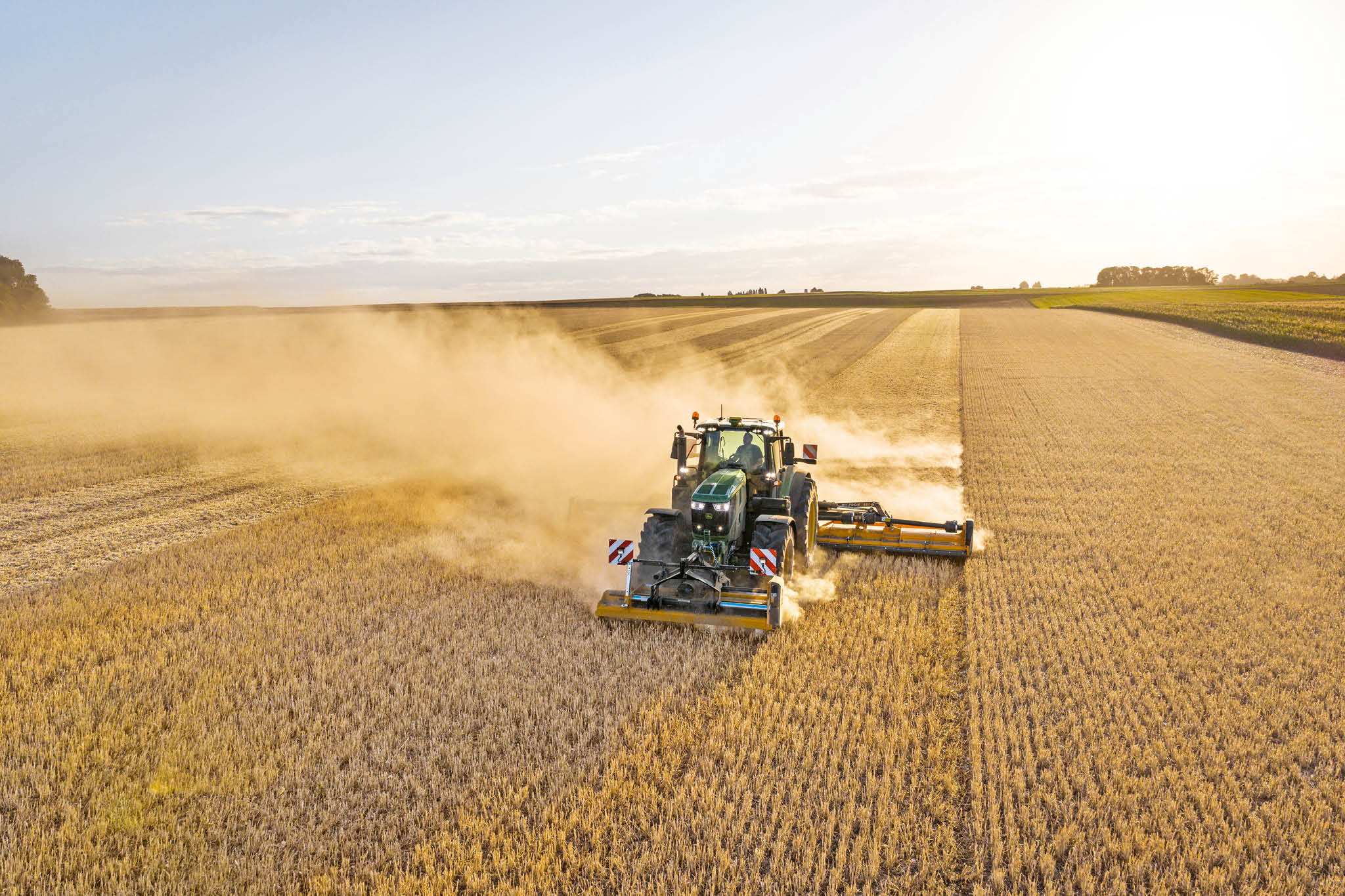Traktor John Deere radu 6M ťahajúci cisternu na hnojovicu pozdĺž vidieckej cesty lemovanej trávou a poľami.