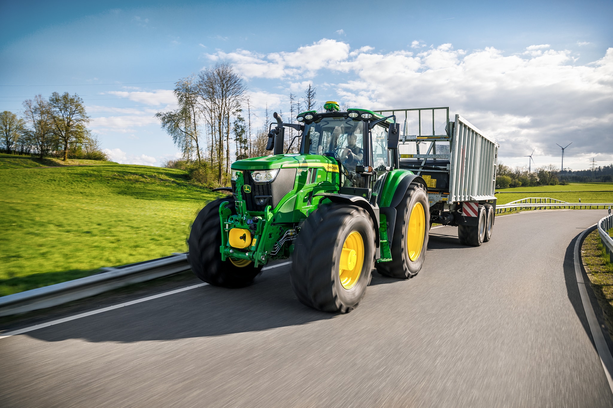 Traktor John Deere radu 6M prepravujúci príves s vysokými bočnicami po spevnenej vidieckej ceste.