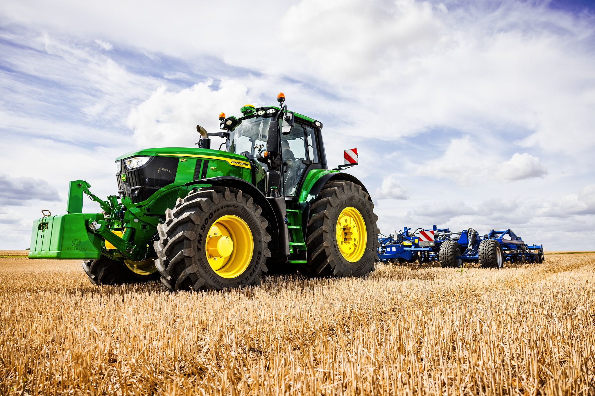 Traktor John Deere radu 6M pri ťahaní obrábacieho príslušenstva po strnisku pod zamračenou oblohou.