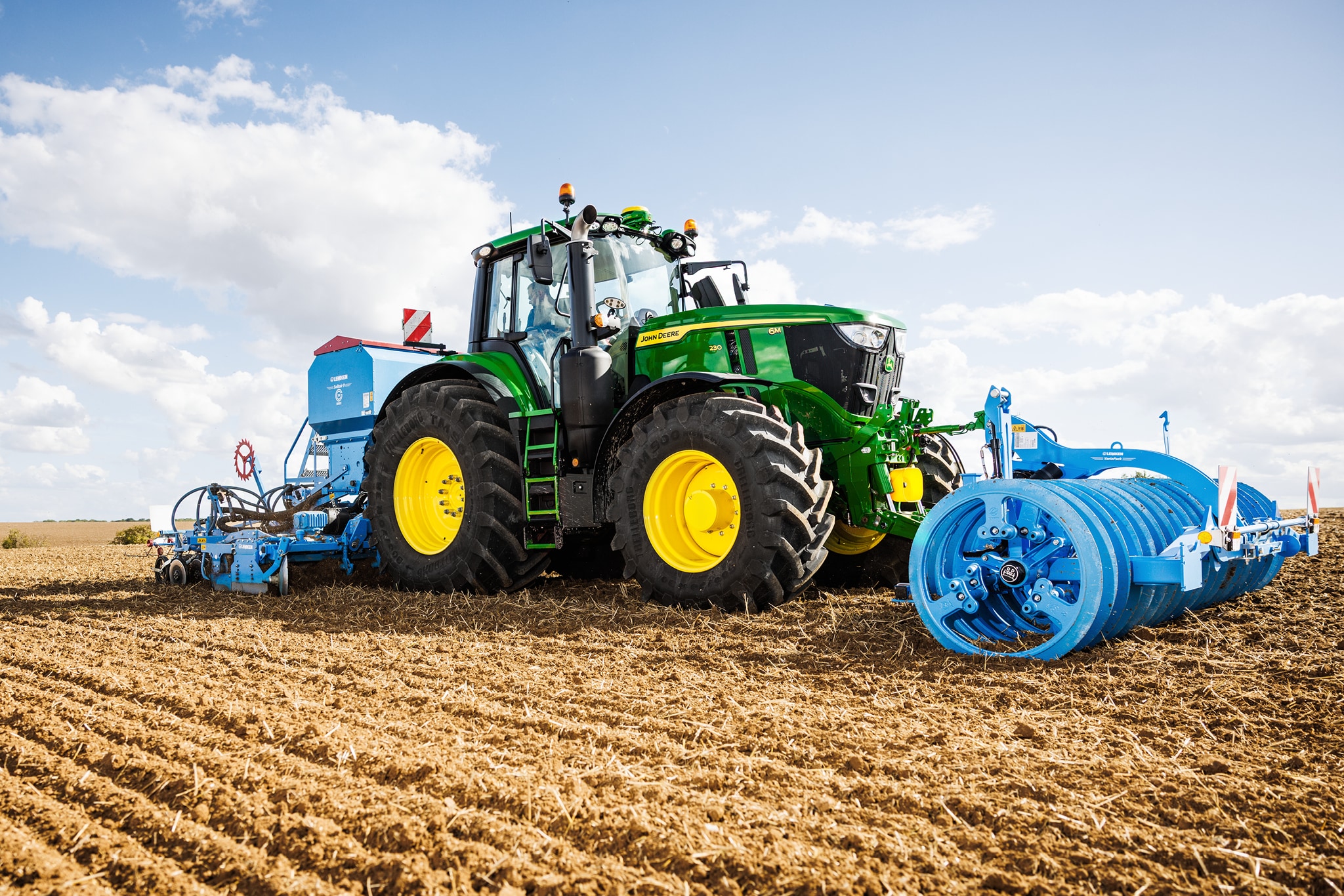 Traktor John Deere radu 6M obrába pole s pripojeným veľkým modrým valcom.