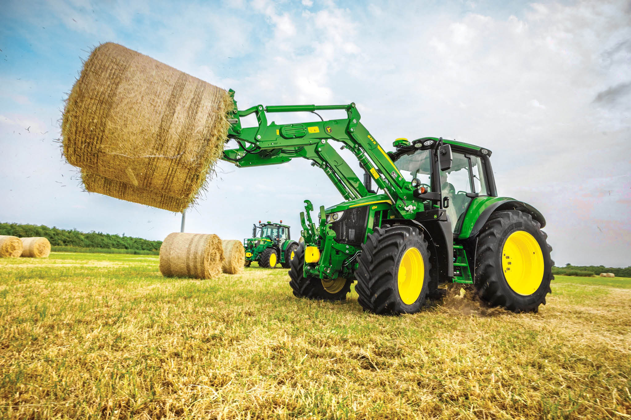 Traktor John Deere radu 6M zdvíha veľký okrúhly balík sena pomocou čelného nakladača na širokom poli.
