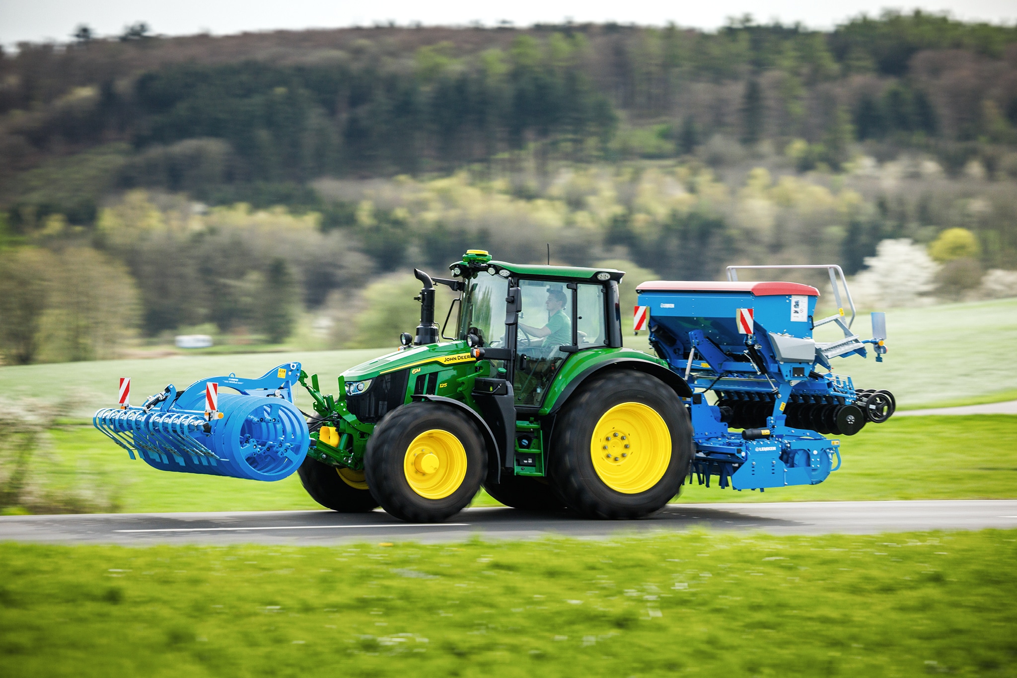 Traktor John Deere radu 6M prepravujúci vpredu namontované príslušenstvo a zadnú sejačku po vidieckej ceste.