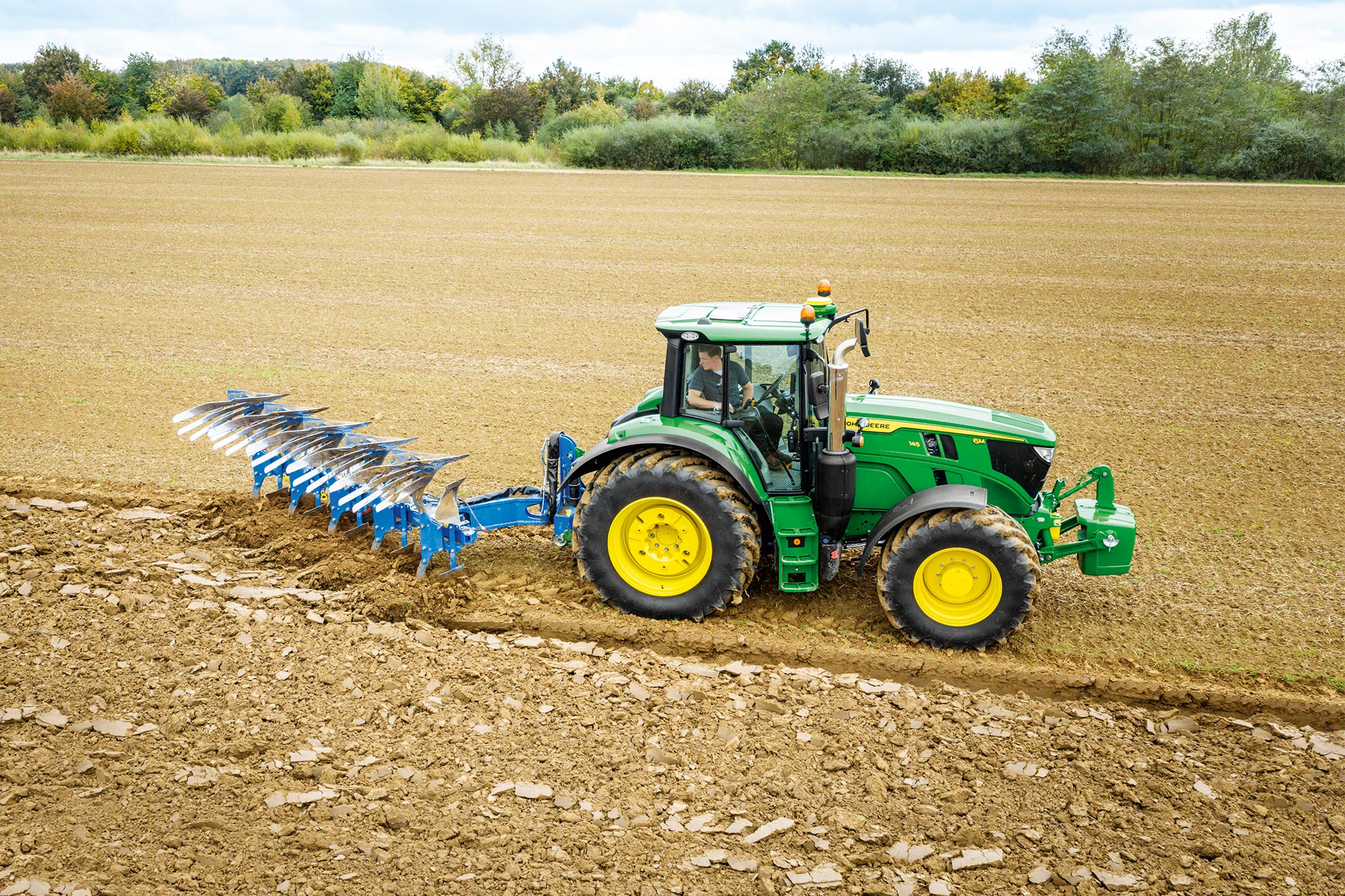 Traktor John Deere radu 6M pracuje na oranom poli so zadným obrábacím príslušenstvom pod zamračeným nebom.