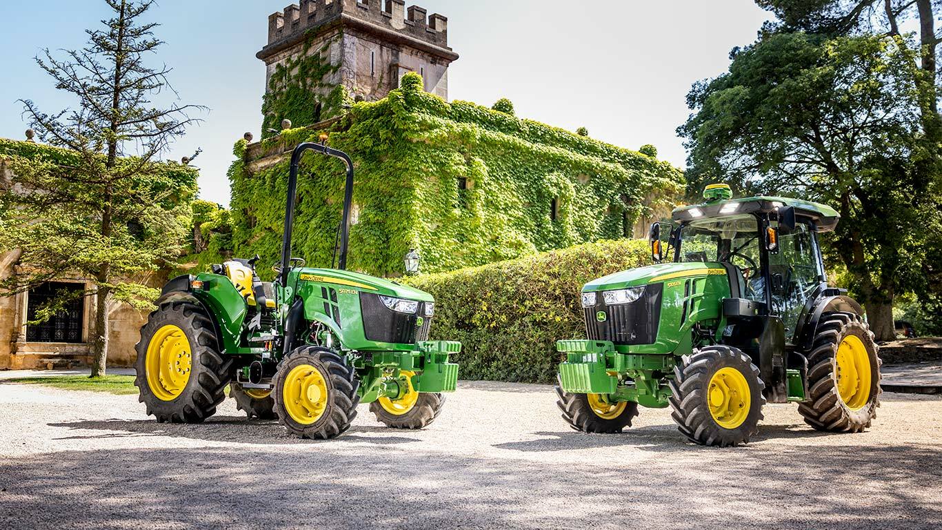 Zelenožltý traktor John Deere 5075EN, ktorý prechádza ovocným sadom po nespevnenej ceste obkolesený ovocnými stromami a zaliaty slnečným svetlom.
