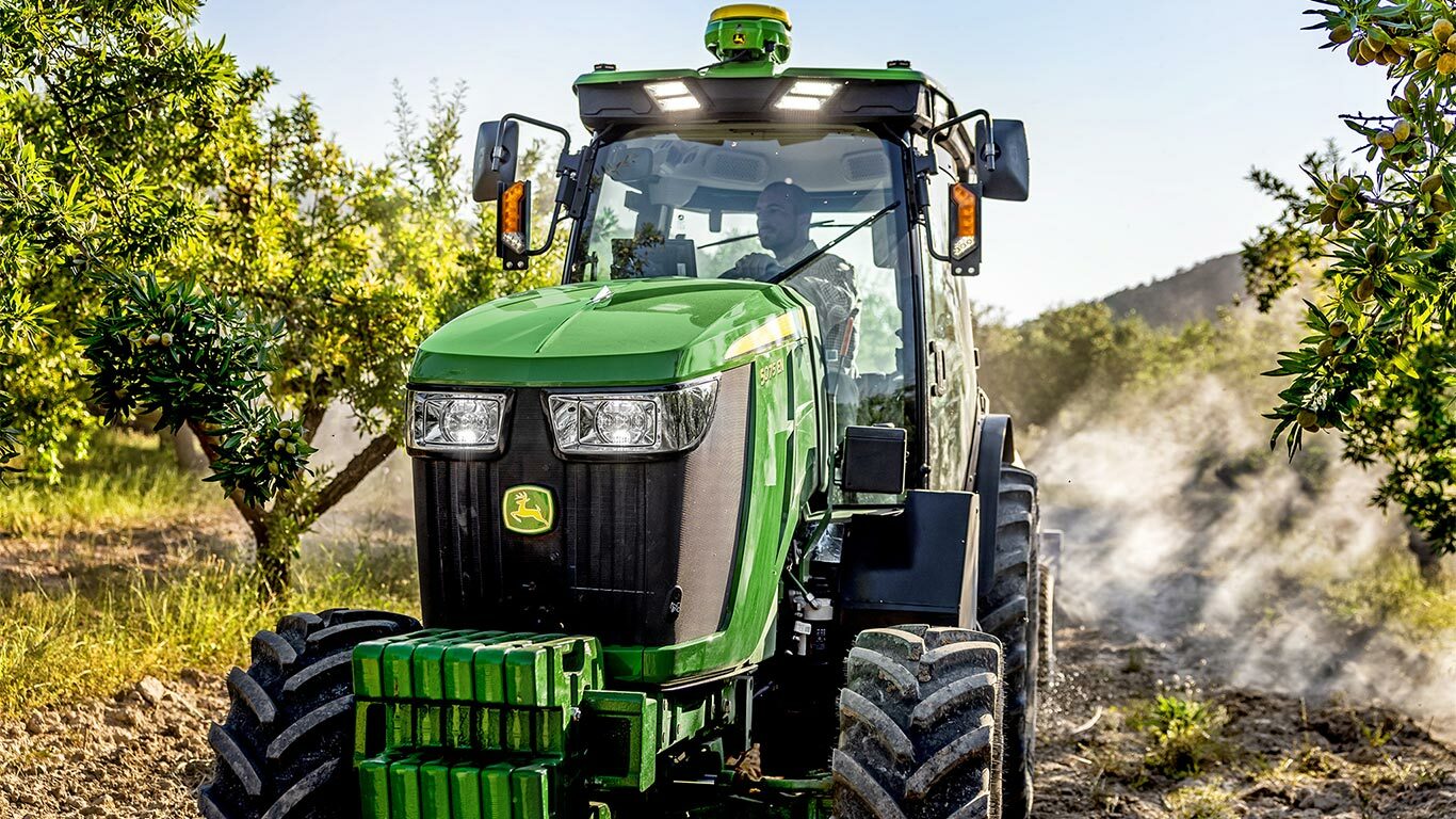 Zelenožltý traktor John Deere 5075EN, ktorý prechádza ovocným sadom po nespevnenej ceste obkolesený ovocnými stromami a zaliaty slnečným svetlom.