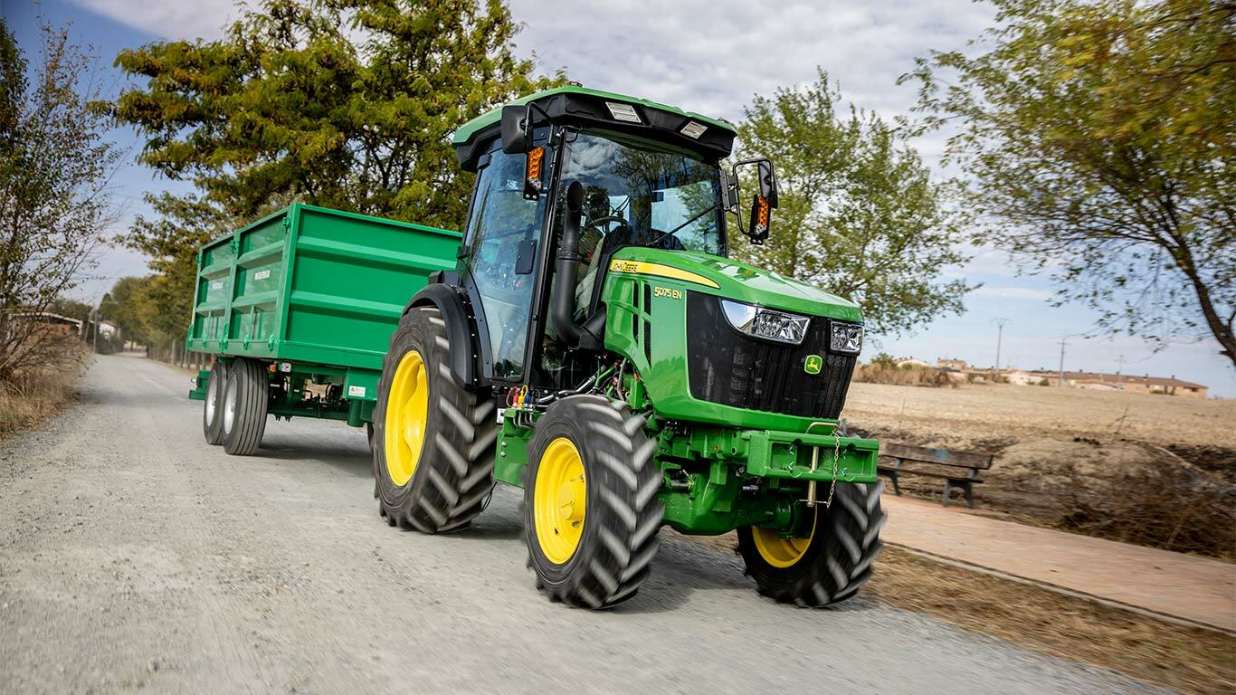 Traktor John Deere 5075EN ťahá zelený príves po vidieckej ceste s stromami v pozadí.