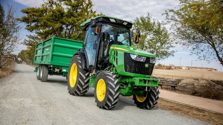 Traktor John Deere 5075EN ťahá zelený príves po vidieckej ceste s stromami v pozadí.