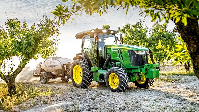 Zelenožltý traktor John Deere 5090EN ťahajúci postrekovač cez ovocný sad počas slnečného dňa.