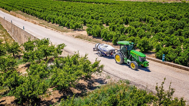 Zelenožltý traktor John Deere 5090EN ťahajúci postrekovač po spevnenej ceste medzi veľkými radmi ovocných sadov pod čírou modrou oblohou.