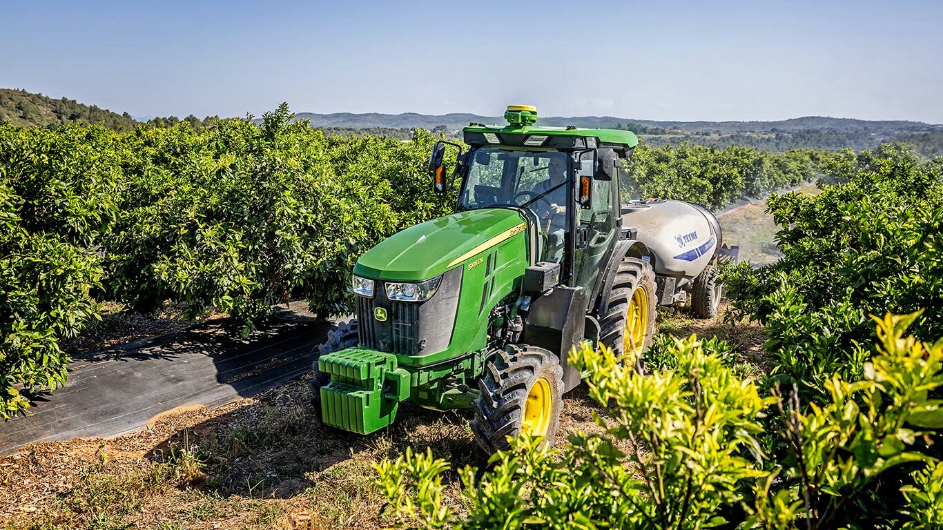 Zelenožltý traktor John Deere 5105EN ťahajúci postrekovač v krásnom ovocnom sade so zvlnenými kopcami v pozadí.