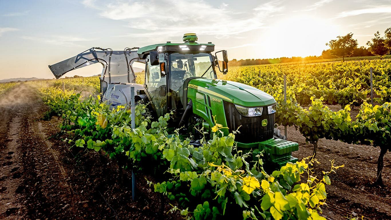 Zelenožltý traktor John Deere 5105EN pracujúci vo vinici pri západe slnka, ktorý postrekuje plodiny medzi radmi viniča.