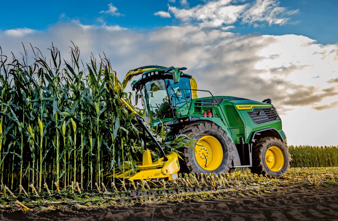 Žací stroj John Deere na zber krmovín na poli pod modrou oblohou s mrakmi.