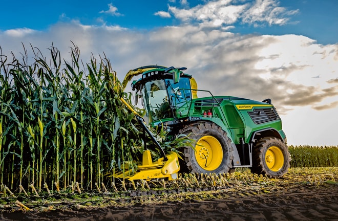 Žací stroj John Deere na zber krmovín na poli pod modrou oblohou s mrakmi.