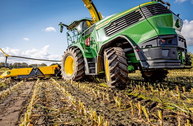 Žací stroj John Deere na zber krmovín pracujúci na kukuričnom poli, na ktorom sú vidieť veľké pneumatiky a rezací mechanizmus stroja na pozadí jasnej modrej oblohy.