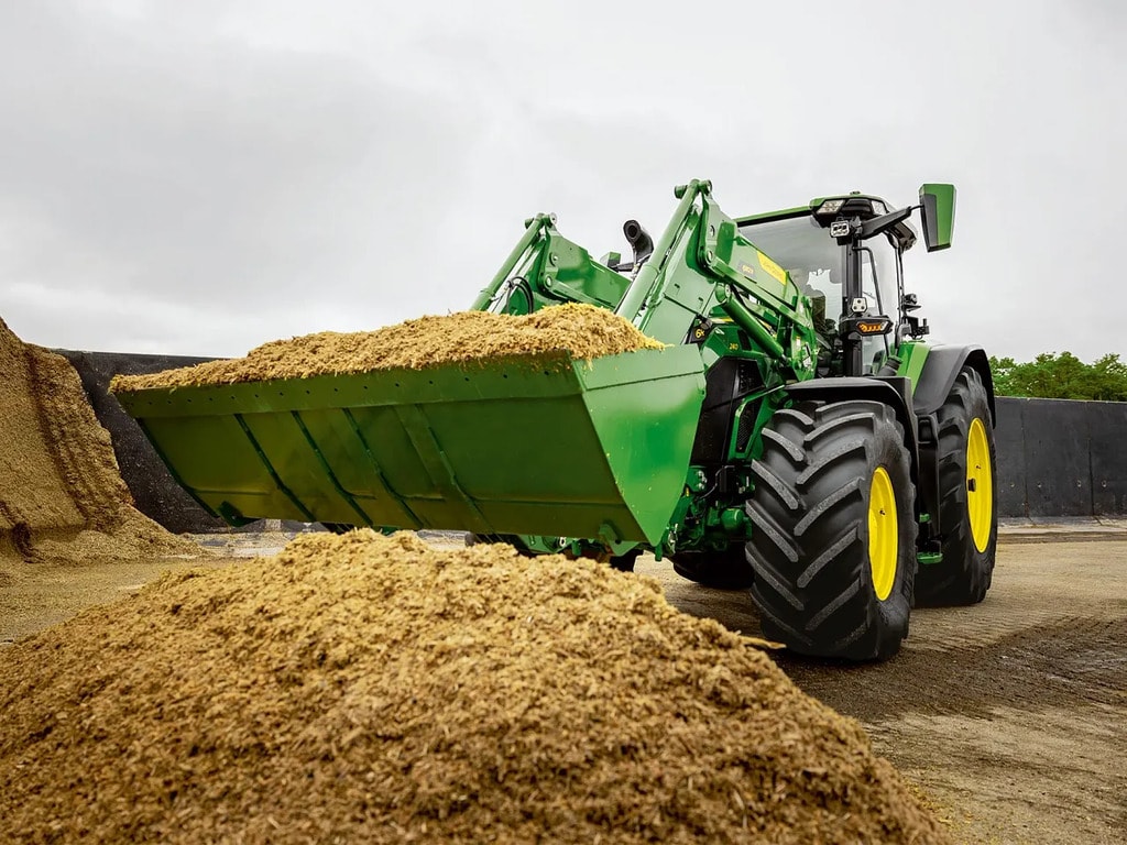 Traktor John Deere prepravuje nasekanú siláž pomocou čelného nakladača v poľnohospodárskom skladovacom priestore.
