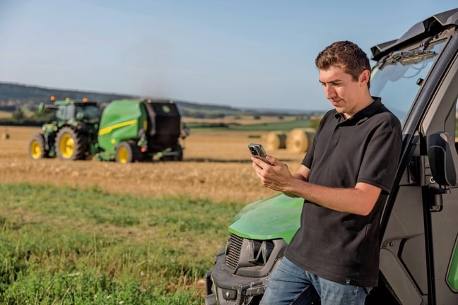 Osoba kontrolujúca údaje dokumentácie balíka v mobilnom telefóne, pričom v pozadí je lis John Deere VR