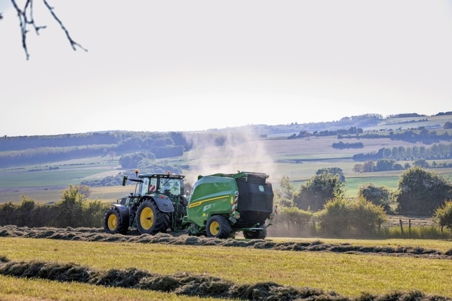 John Deere V462R ťahaný traktorom John Deere 6R 185, ktorý lisuje seno
